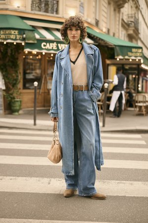 Moteris su garbanotais plaukais, vilkinti ilgą džinsinį paltą, plačiomis kelnėmis, šviesiai rudą palaidinę ir žaliais auskarais, stovi gatvės pėsčiųjų perėjoje, laikydama šviesiai rudą rankinę.