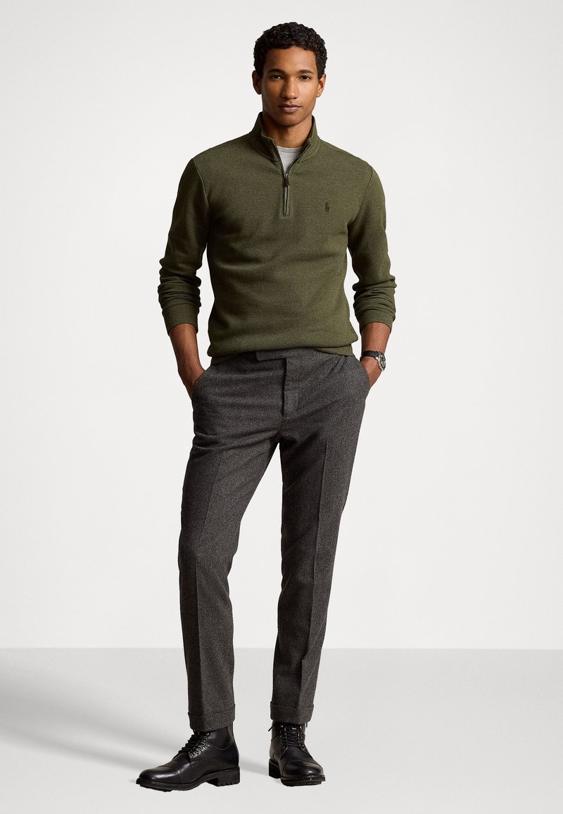 Olivgrüner Pullover mit Reißverschlusskragen, Wollmischung, mit subtiler Logostickerei. Graue, maßgeschneiderte Hose aus strukturiertem Stoff und schwarze Schnürlederschuhe.
