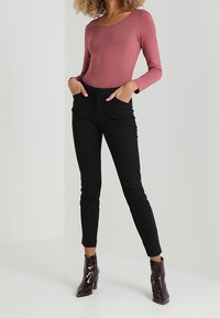 Långärmad topp med rundad halsringning i dammrosa, kombinerad med svarta, figurnära byxor. Modellen bär blanka, mörkt burgunderfärgade ankelstövlar.