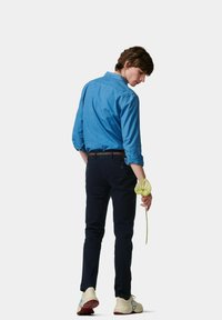 Lichtblauwe denimshirt, donkere slim-fit broek, witte sneakers met kleuraccenten, houdt een groene bloem vast, met rug naar de camera, neutrale achtergrond.