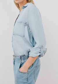 Camicetta di denim azzurro chiaro con maniche lunghe, polsini arrotolati e tasca frontale; abbinata a pantaloni corti con cintura elastica azzurro chiaro.