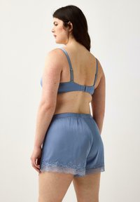 Modella di spalle, indossa un reggiseno blu e pantaloni satinati abbinati con bordo in pizzo. Lo sfondo semplice mette in risalto il design dell'outfit.