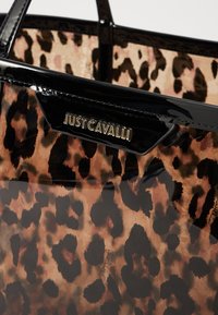 Bolso transparente con un diseño de estampado de leopardo marrón y negro, acentos negros brillantes y una placa con el logo dorado. Textura suave.