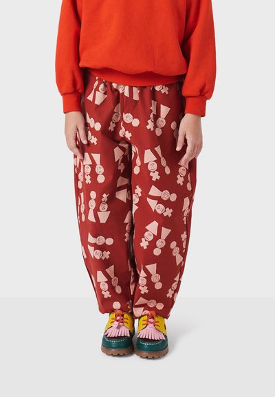 Červené nohavice s abstraktnými tvarmi a tvárami, s voľným strihom; skombinované s mnohofarebnými topánkami a oranžovým sveterom.