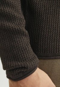 Tamno smeđi pleteni džemper izbliza, s teksturiranim uzorkom, rebrastim manžetama i glatkim područjem ispod pazuha.