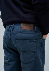 Temno modri kavbojki iz denima z rjavim usnjenim blagovno znamko "SALSA JEANS". Oblikovanje vključuje dve zadnji žepki z vidnim šivanjem.