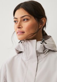 Hellgraue wasserdichte Jacke mit hohem Kragen, elastischen Zugdetails und glatter Textur, geeignet für verschiedene Wetterbedingungen.