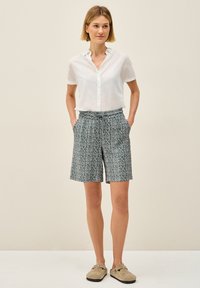 Camisa blanca ligera de botones, pantalones cortos azules con un diseño geométrico, dos bolsillos laterales y zapatos bajos beige casuales.