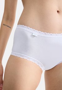 Culotte en coton blanc avec bordure en dentelle à la taille, présentant une texture lisse et une coupe ajustée. Petite étiquette visible sur le côté.