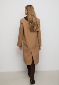 Cappotto di lana color cammello con silhouette dritta, maniche lunghe e spacco sulla schiena. Indossato sopra stivali marroni fino al ginocchio.