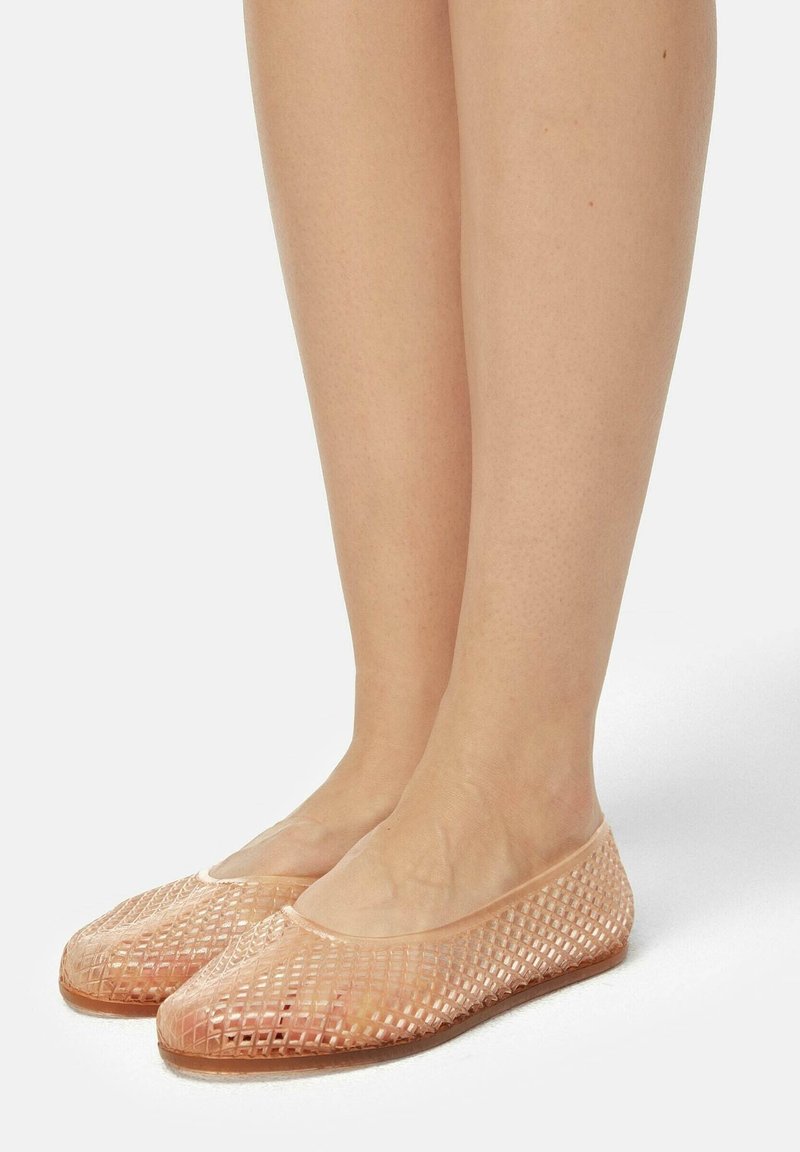 Jambes portant des ballerines beiges translucides avec un motif en treillis, debout sur une surface blanche.