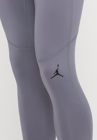 Vysoké a priliehavé legíny z svetlo šedej tkaniny s tmavo šedým prešívaním a čiernym logom Jumpman na ľavom stehne.