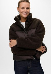 Lächelnde Frau trägt eine dunkelbraune Fleecejacke mit gesteppten Abschnitten, dunkle Jeans und silberne Hängeohrringe vor einem schlichten weißen Hintergrund.