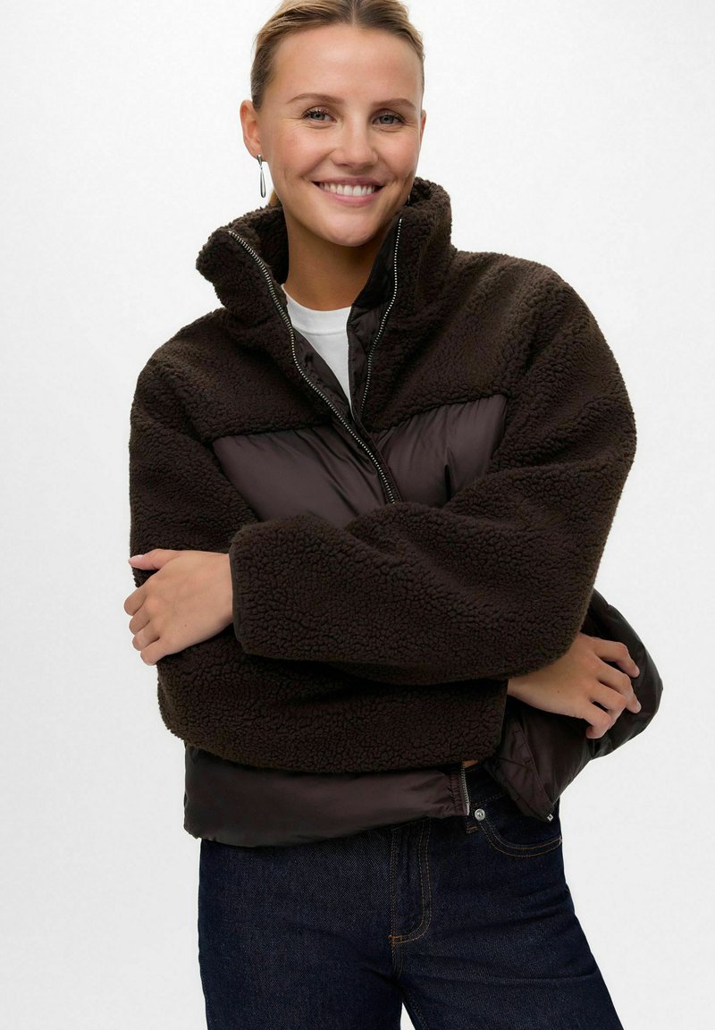 Lächelnde Frau trägt eine dunkelbraune Fleecejacke mit gesteppten Abschnitten, dunkle Jeans und silberne Hängeohrringe vor einem schlichten weißen Hintergrund.