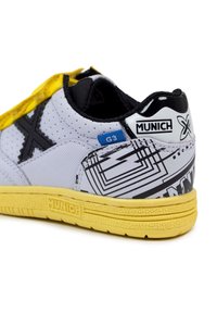 Zapatilla blanca y negra con una superficie texturizada, acentos amarillos, patrones geométricos y una lengüeta trasera negra con la marca "MUNICH".