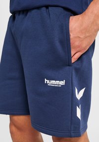 Man som bär marinblå sportshorts med vit "hummel"-logotyp och chevrondesign, handen vilar i högra fickan.