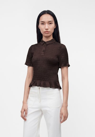 Jeune femme aux longs cheveux noirs portant un chemisier texturé marron foncé à manches courtes et un pantalon blanc taille haute, debout devant un fond blanc.