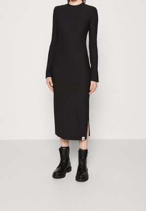 Femme portant une robe midi noire à manches longues avec une fente sur le côté, associée à des bottines de combat noires à lacets, debout devant un fond uni.