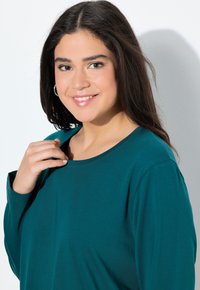 Teal langslip shirt met een ronde hals, gemaakt van zacht materiaal. Heeft een comfortabele, relaxte pasvorm en licht afgezakte schouders.