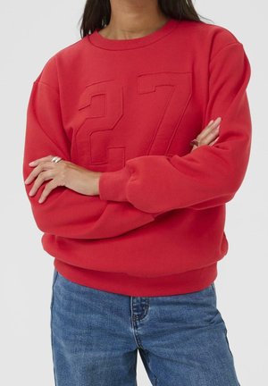 Persona con una sudadera roja con el número 27 en relieve y vaqueros azules, con los brazos cruzados, sobre un fondo claro y liso.