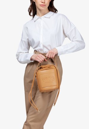 Tanskleurige leren trekkoordtas met een gegraveerd logo. Vierkante vorm, gladde textuur en verstelbare banden. Draagt met een wit shirt en beige broek.