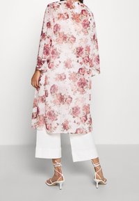 Žena v průhledném květinovém kimonu s růžovými růžemi, oblečená v bílých širokých kalhotách a bílých sandalech na podpatku, stojící na jednobarevném pozadí.