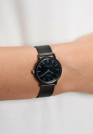 Sort minimalistisk armbåndsur med mesh-rem båret på venstre håndled, viser tidspunktet cirka 2:20 mod en lys neutral baggrund.