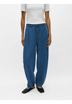 Person trägt weite blaue Jeanshosen mit elastischen Bündchen, weißes kurzärmeliges Hemd, schwarze flache Schuhe mit quadratischer Spitze und ein Armband am rechten Handgelenk.
