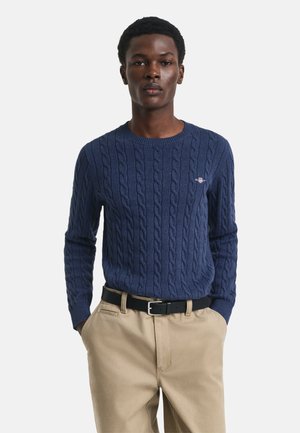 Dunkelblauer Kabelstrickpullover mit rundem Halsauschnitt, der ein kleines Logo-Emblem trägt, kombiniert mit hellen Khakihosen und einem schwarzen Gürtel.
