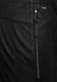 Svarta byxor i faux läder med en blixtlås-ficka med silverdetaljer, slät textur och minimala sömnadsdetaljer.