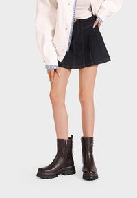 Stivaletti in pelle marrone con finitura liscia, cerniera laterale, dettagli a coste e una spessa suola in gomma nera. Indossati con una gonna di jeans.