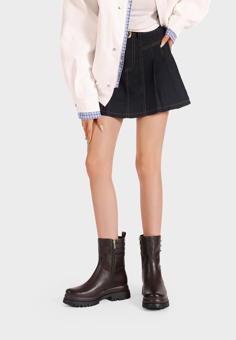 Stivaletti in pelle marrone con finitura liscia, cerniera laterale, dettagli a coste e una spessa suola in gomma nera. Indossati con una gonna di jeans.