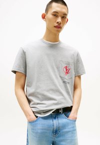 Camiseta de algodón gris con cuello redondo, mangas cortas, un logotipo de bolsillo rojo y un corte recto, combinada con jeans de mezclilla azul claro.
