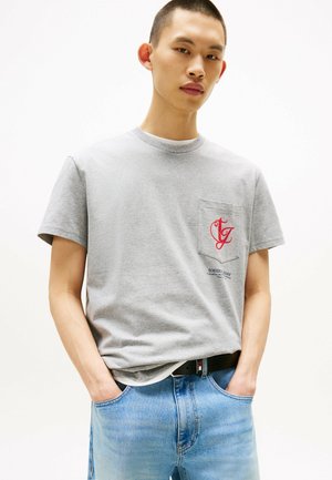 Camiseta de algodón gris con cuello redondo, mangas cortas, un logotipo de bolsillo rojo y un corte recto, combinada con jeans de mezclilla azul claro.