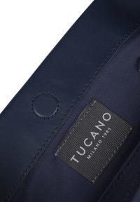 Interieur van een marineblauwe leren tas met een label met de tekst "TUCANO MILANO 1985." Bevat een ronde drukknop en een gestructureerde voering.