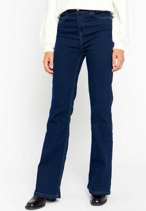 Personne portant un jean bootcut bleu foncé, des chaussures noires et un haut blanc à manches longues, debout devant un fond blanc uni.
