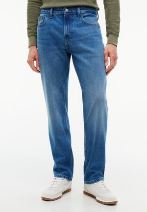 Homme portant un jean bleu moyen à jambes droites, des baskets blanches et une chemise à manches longues vert olive, debout devant un fond blanc.