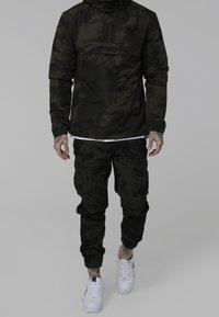 Mand iført sort camouflagejakke og cargo-bukser med hvide sneakers mod en ensfarvet grå baggrund.