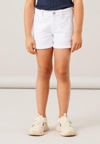 Shorts in denim bianco con taglio classico, dotati di tasche frontali e bottone in metallo. Abbinati a scarpe da ginnastica beige con cinturini.