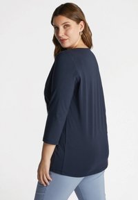 Tummansininen tunika, jossa on kolme neljäsosaa hihoista, pehmeä kangas ja pyöreä kaula-aukko, puettuna vaaleansinisten housujen päälle. Yksinkertainen ja elegantti muotoilu.