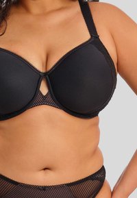Reggiseno nero a coppa morbida con rifinitura in pizzo, dotato di supporto con ferretto e una sezione in rete al centro. Spalline regolabili e chiusura con gancio.