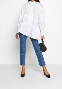 Bílá košile s krátkým rukávem a nabíranými švy, zkombinovaná s přiléhavými modrými džíny a černými lodičkami s špičatou špičkou, držící kulatou černou tašku.