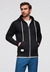 Felpa nera con zip, dotata di lacci bianchi, tasche frontali e polsini a coste. Indossata su una maglietta bianca con motivo e abbinata a jeans blu.