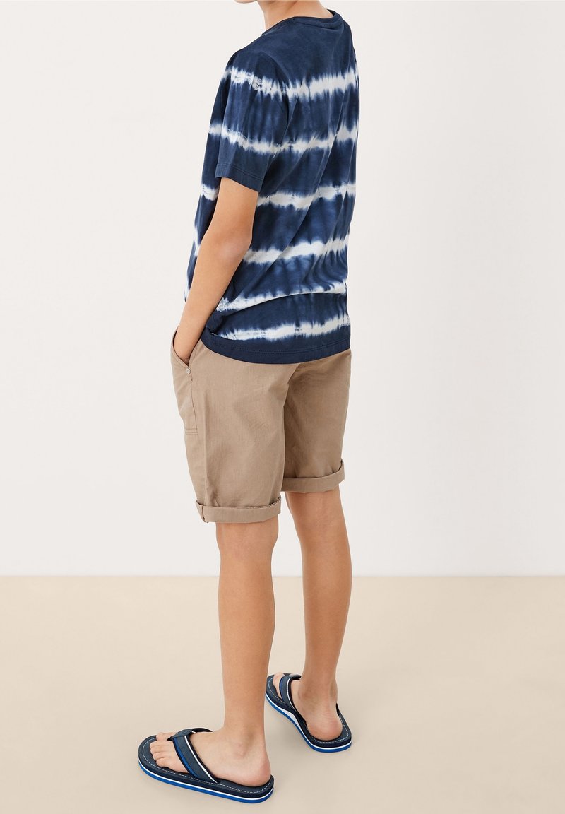 T-shirt tie-dye bleu marine et blanc, shorts beige roulés et tongs bleues. La tenue présente une coupe décontractée et des matériaux confortables.