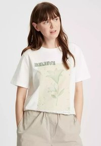 Biała bawełniana koszulka z krótkim rękawem, zdobiona pastelowym wzorem kwiatowym oraz napisem „BELIEVE” w kolorze zielonym na górze. Luźny krój.