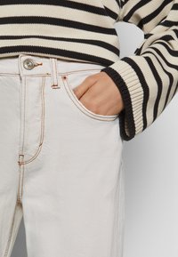 Jeans de ganga brancos com costura laranja visível, apresentando um bolso frontal e um botão prateado. A parte de cima é um suéter listrado em preto e creme.