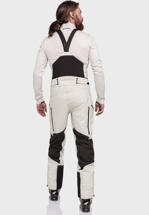 Mann mit langen Haaren und Bart, der mit dem Rücken zur Kamera steht, trägt weiße und schwarze Skihosen mit Hosenträgern und schwarze Skischuhe.