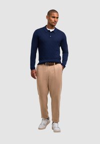 Pullover lavorato a maglia in navy con motivo a rombi, pantaloni beige su misura e sneakers bianche con accenti neri. Outfit casual su sfondo grigio.
