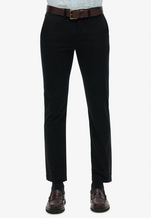 Pantalones negros con un corte ajustado, que cuentan con un cinturón de cuero marrón y una parte delantera plisada. Llevados con calcetines oscuros y mocasines marrones.