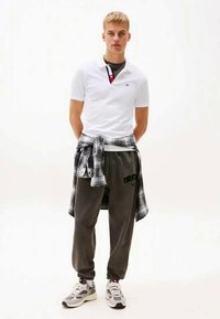 Giovane in piedi con le mani dietro la schiena, indossa una polo bianca, una camicia a quadri nera annodata in vita, pantaloni jogger scuri e sneaker bianche.
